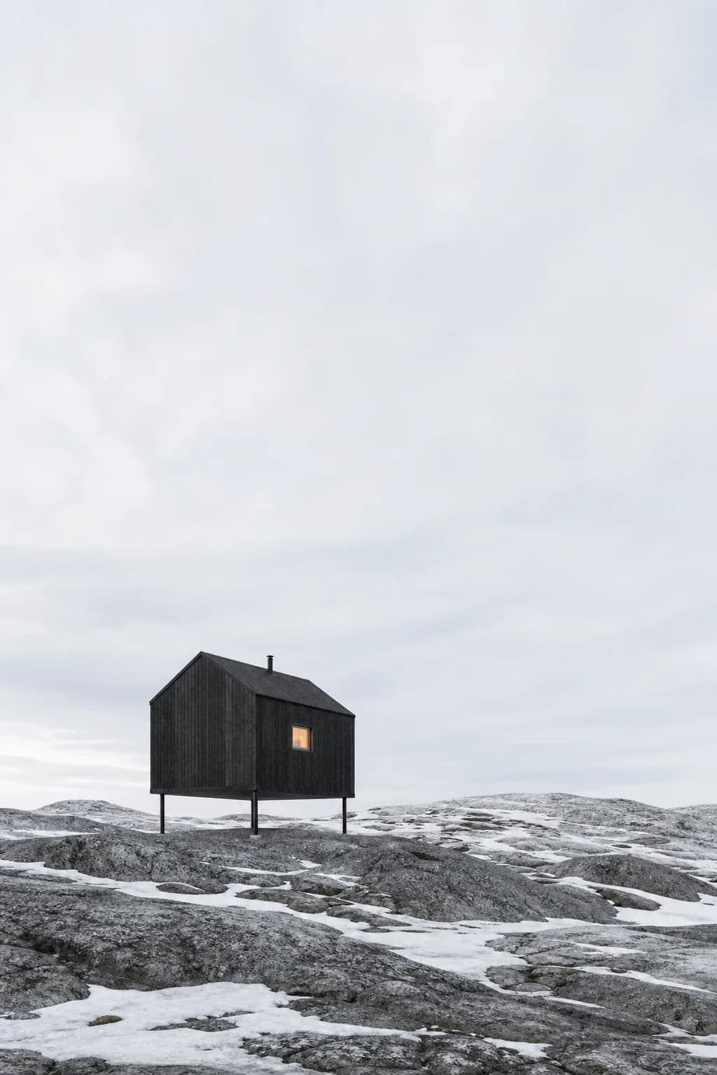 Fjellrev Cabin: a small dark-stained timber cabin raised on four slender steel legs above bare bedrock, one warm window lit under a vast pale sky