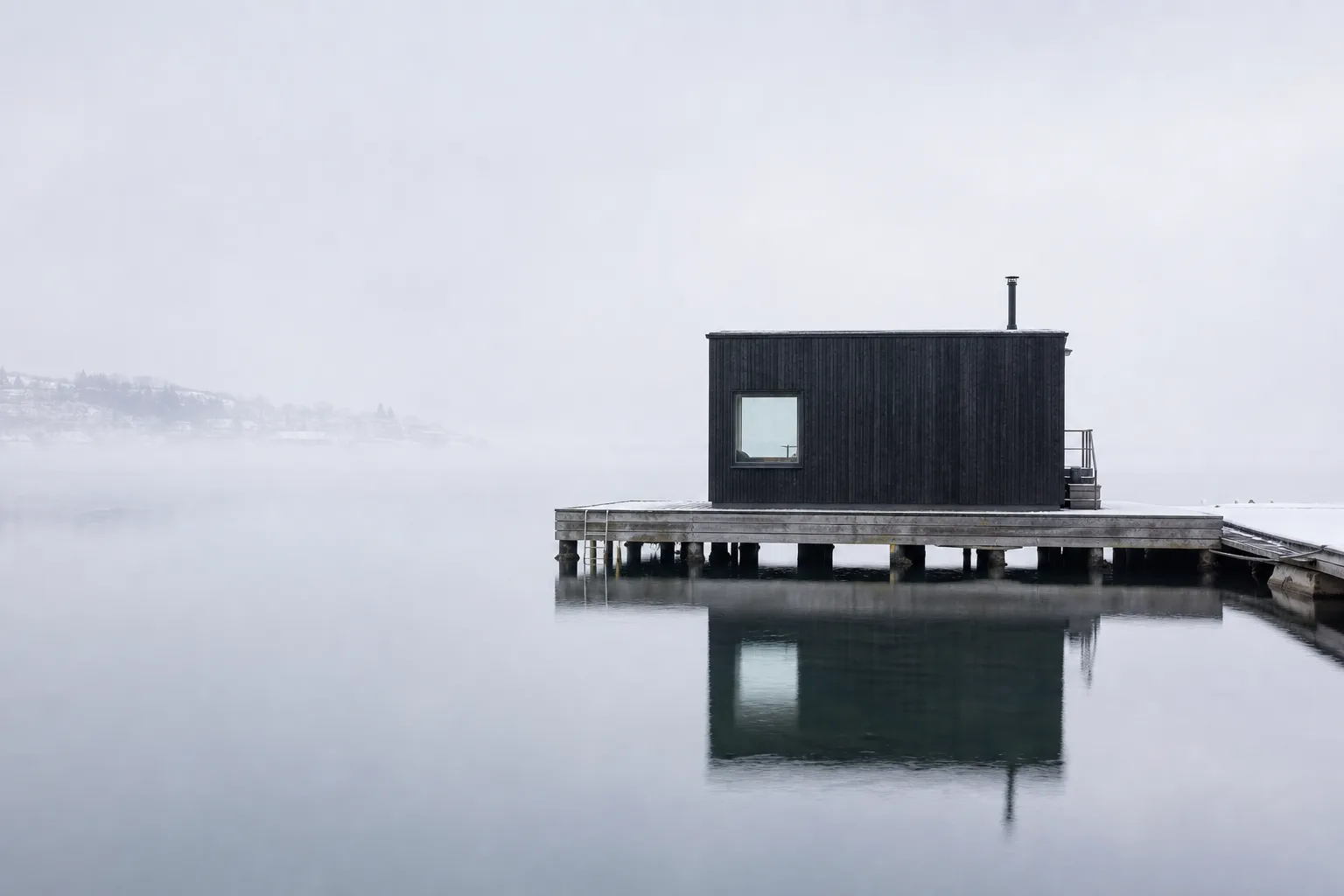 Sørenga Harbour Sauna: a charred black timber volume on a wooden pier over still grey harbour water, reflected in the fjord with winter mist behind