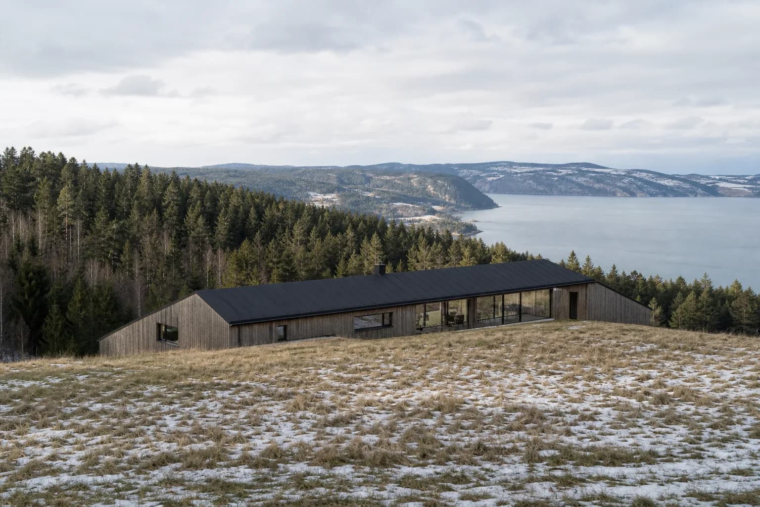 Vindsletta House: a long, low timber house whose dark roof follows the crest of a hill, pine forest behind and a pale fjord beyond