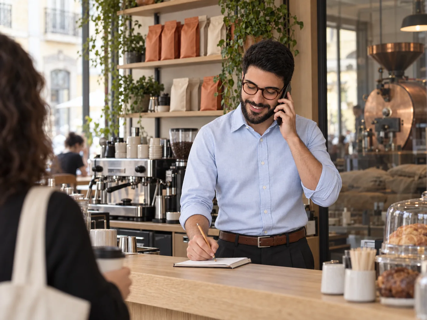 The owner of a small coffee shop behind the counter on a busy morning, the phone at his ear, writing an order into a paper notebook while a customer waits.