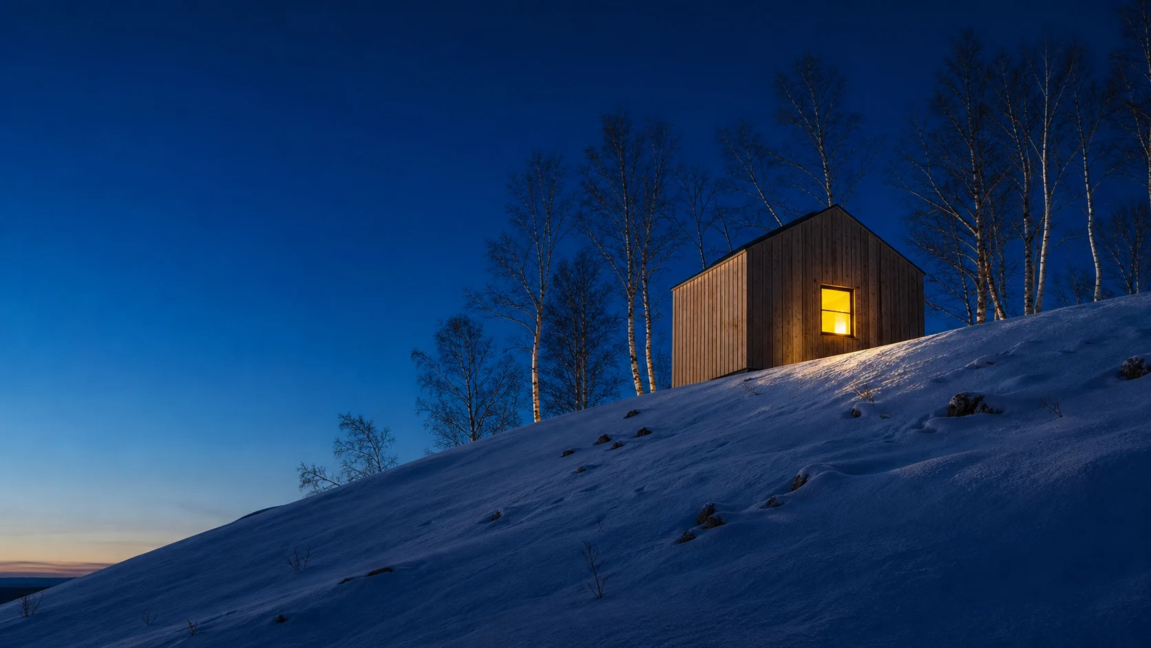 A small timber annexe at blue hour, its one window lit warm, on the crest of a snowy birch slope