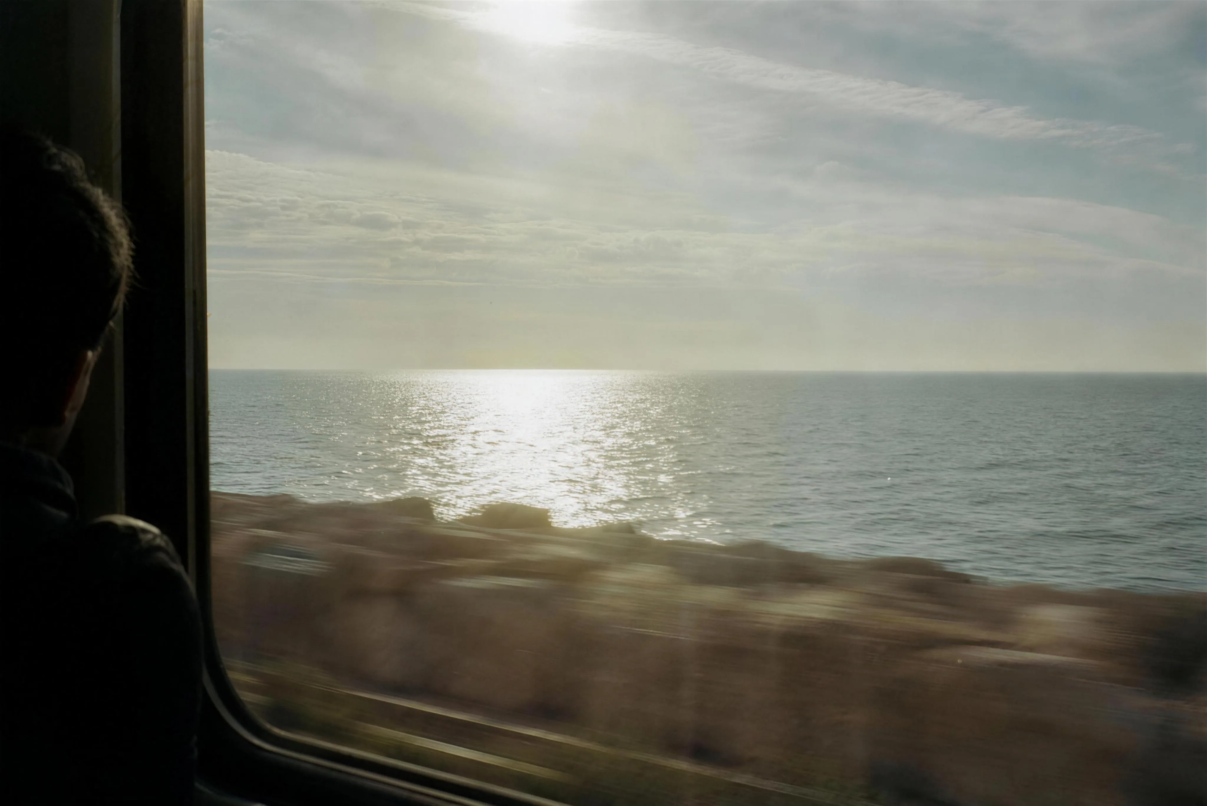 The flat winter Mediterranean through the window of a moving train, silver glare off the sea, the silhouetted head of a single passenger at the frame's edge