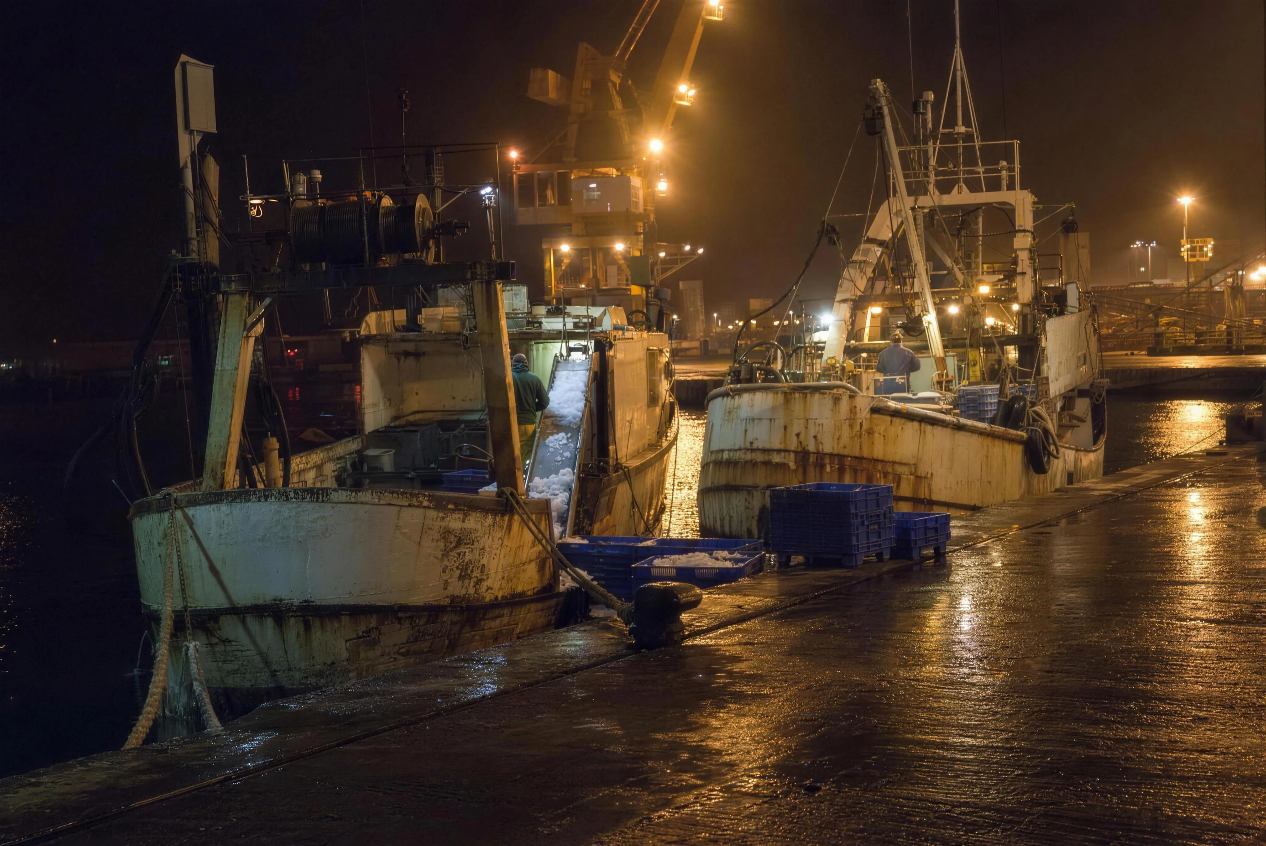 Two rusty trawlers moored side by side at a wet quay at night, ice sliding into blue crates under sodium lamps, a gantry crane in the amber haze behind