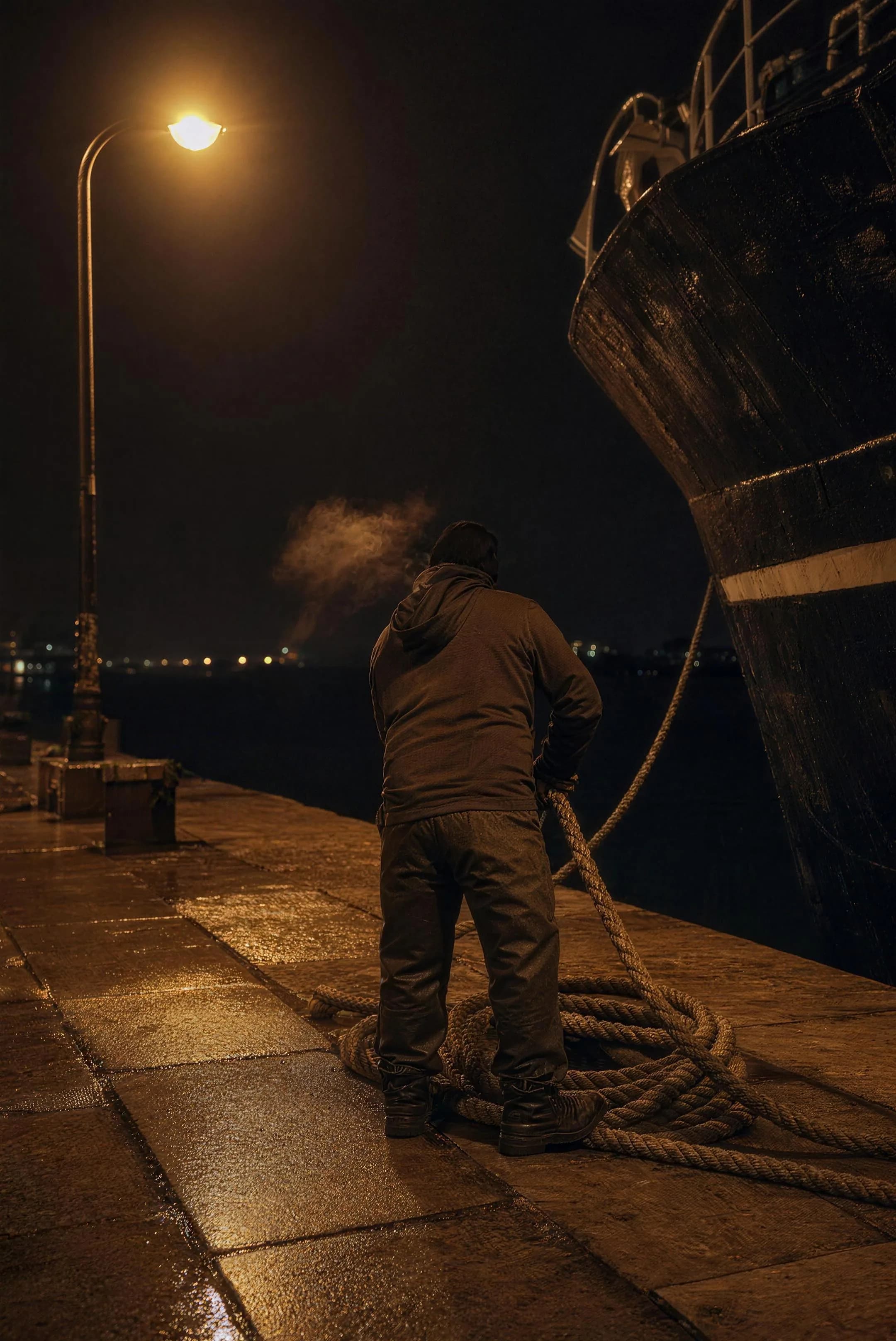 A dockworker seen from behind coiling heavy rope under a single sodium lamp, his breath visible, a trawler's dark bow towering at the edge of the frame