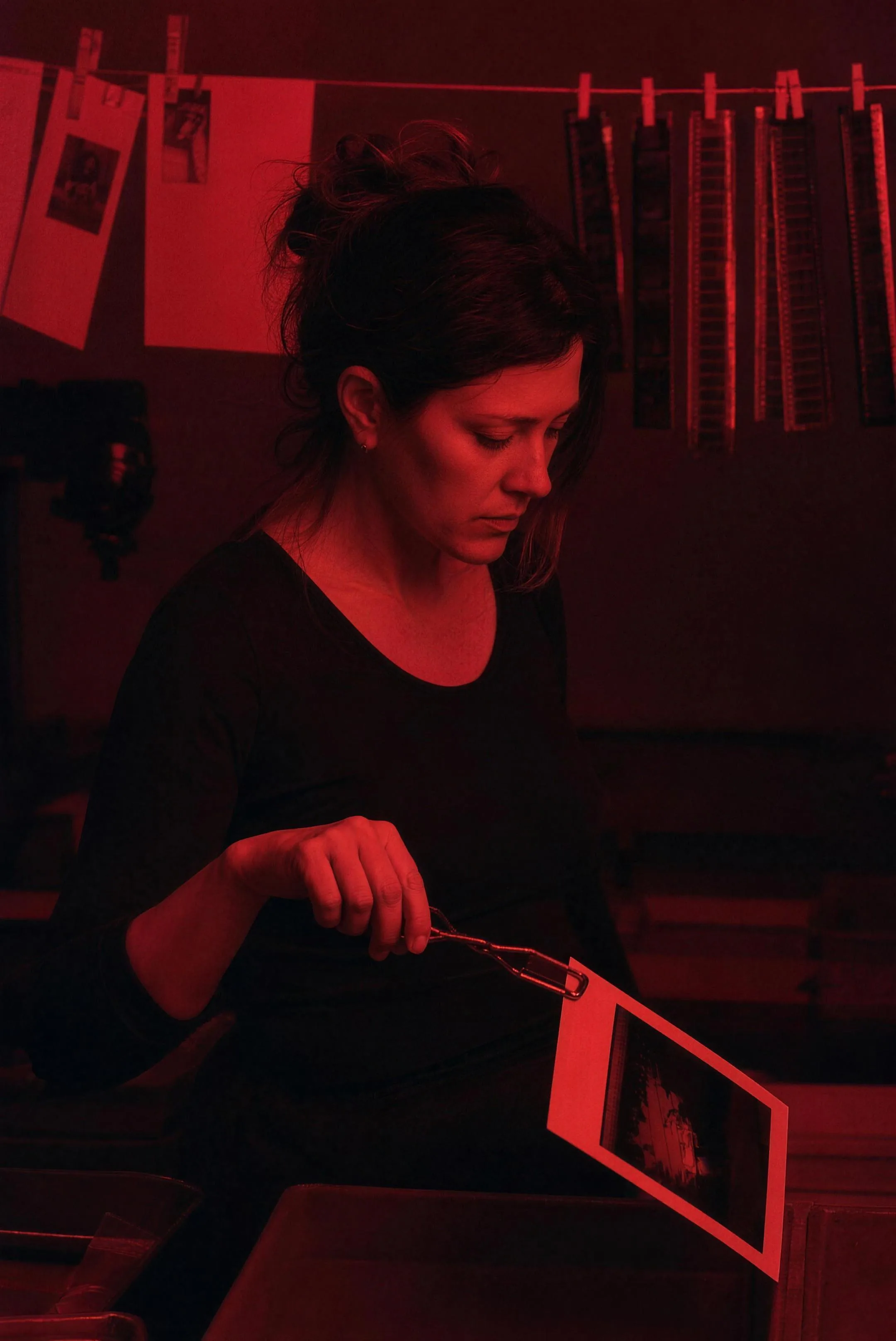 Mara Lindqvist in her darkroom under the red safelight, looking down at a freshly developed print held in tongs, negatives hanging behind her