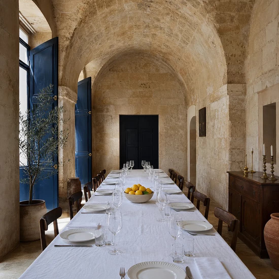 The long dining table under the stone barrel vault, laid in white linen with one bowl of lemons, empty and set for service