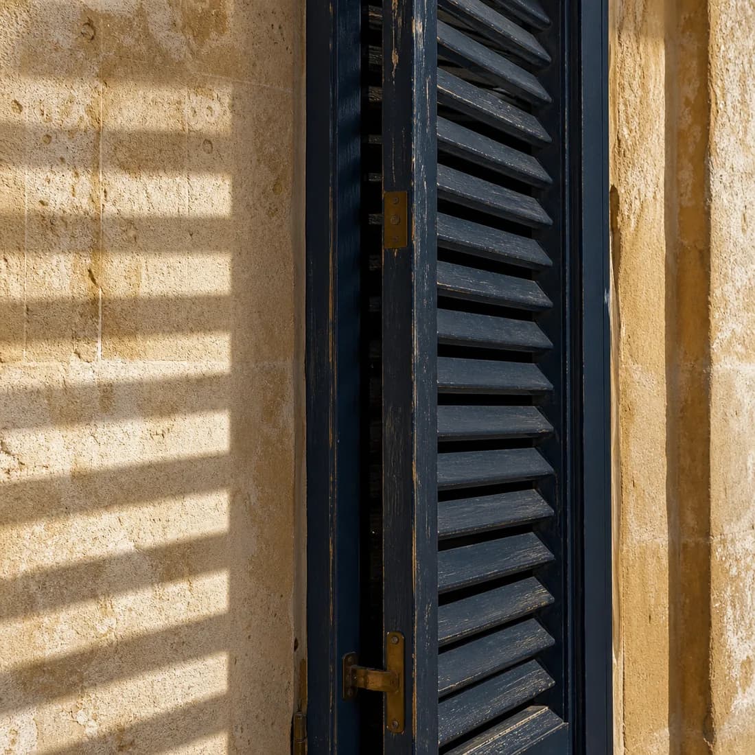 A tall ink-blue louvred shutter, half closed, against a wall of honey limestone striped by hard Sicilian sun