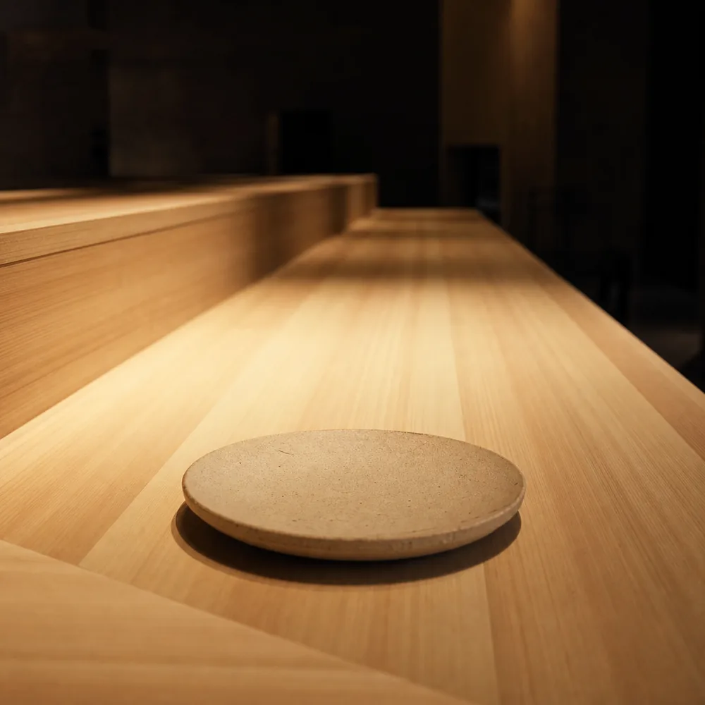 The empty hinoki counter seen from a diner's seat, a warm pool of light on the wood and the room soft beyond it