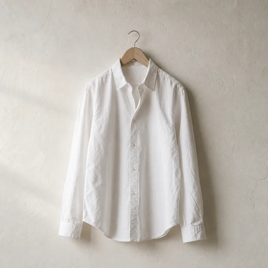 A white cotton shirt on a plain wooden hanger against a bare plaster wall, raking light across the cloth