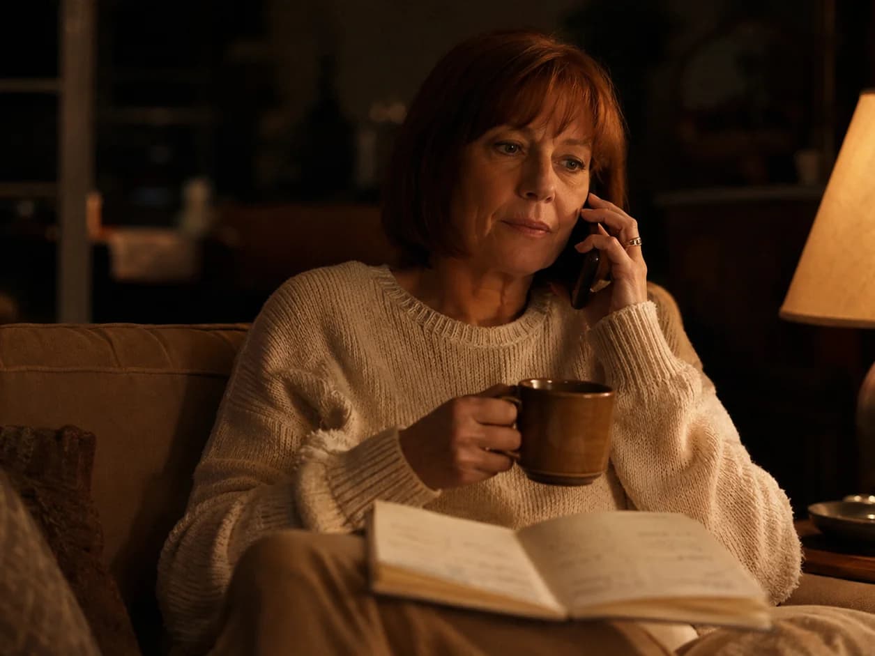 A woman on her sofa late in the evening under one lamp, phone to her ear listening to a recorded message, her own diary open on her knee with the free evenings marked