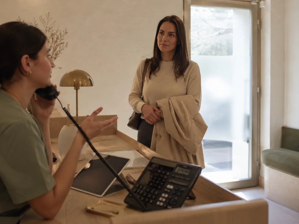 The receptionist on the phone with a hand over the receiver, explaining a price again, while a new client stands at the desk holding her coat and waiting