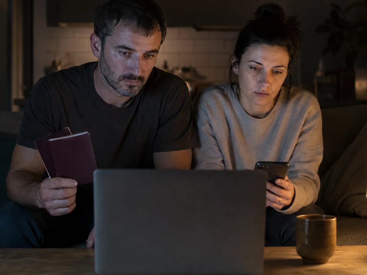 A couple on their sofa at eleven at night with a laptop open on an agency's contact page, one holding two passports and the other checking the time on a phone